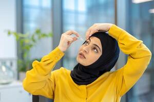A woman in a hijab applies eye drops to her eye, likely to treat dryness or irritation, indoors in a business setting. photo