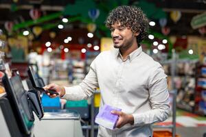 A smiling man uses contactless payment at a grocery store checkout. He holds his phone near the payment terminal and a package of groceries. photo