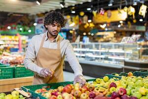 A grocery worker wearing an apron and gloves carefully selects apples from a fresh produce display, checking for quality. photo