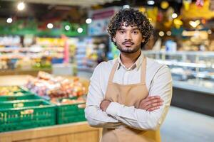 A supermarket employee, wearing an apron, poses with arms crossed while looking directly at the camera. The blurred background suggests a busy store. photo