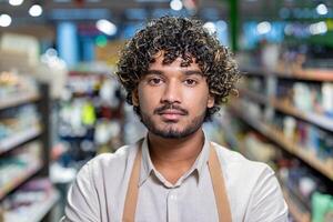A portrait of a store worker standing between shelves with a serious expression. He wears a light shirt and an apron, looking directly at the camera. photo