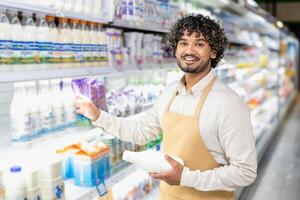 A smiling worker in a supermarket holds a milk bottle, restocking dairy products on the shelf. The focus is on customer service. photo