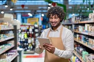 A smiling store clerk checks inventory with a tablet in a grocery store setting. Shelves filled with products surround the employee. photo