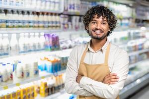 A cheerful store employee stands with folded arms, smiling in front of the dairy section filled with products. The grocery store worker looks confident and ready to assist. photo