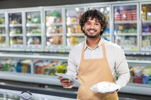A smiling grocery store employee holds a package of food while looking at the camera. He is standing near a freezer. photo