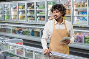 A grocery store employee smiles, using a tablet while checking the refrigerated goods section in the store. He wears an apron. photo