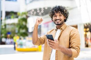 A cheerful man celebrates a success while looking at his smartphone, with a raised fist. He is smiling broadly. The background is blurred, suggesting a public space. photo