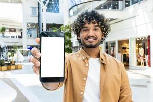 A smiling man with curly hair holds up his smartphone with a blank screen in a shopping mall setting. He is smiling directly at the camera. photo