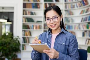 A smiling asian woman with glasses takes notes while wearing earphones, set in a library setting with a backdrop of bookshelves. photo