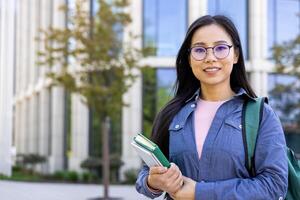 A smiling Asian woman stands in front of a modern building, holding books and a tablet with a backpack. photo