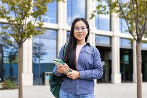 A smiling asian student, wearing glasses, standing in front of a university building with books in hand, carrying a backpack. photo