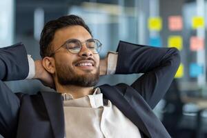 Successful businessman resting while sitting at his desk inside the office. Well done man with his hands behind his head with closed eyes smiling, satisfied with results of achieving the workplace. photo
