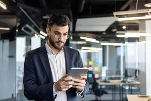 Serious confident businessman with tablet computer. Man in business suit at workplace, using online application, viewing financial investment data, inside office. photo