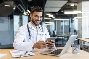 Successful joyful doctor with tablet computer sitting at desk with laptop. Man in white medical coat, consulting patients online, texting and using call. photo