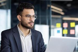 Close-up photo of serious confident businessman at work, thinking man reading electronic report. Businessman at workplace inside office working with computer.