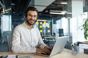 Portrait of young successful company employee with headset. Online customer support consultant smiling and looking at camera, using laptop for call, working inside office. photo