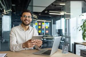 A smiling programmer holding a tablet sits at a desk with a laptop. He looks happy and content in his modern office setting photo