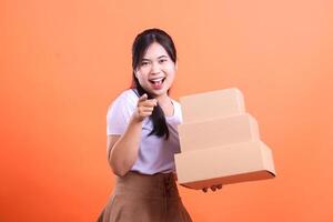 A young woman with a big smile is pointing at the camera while holding a stack of delivery boxes in her hands isolated on orange background. photo