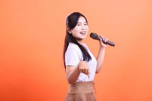A happy young Asian woman with a microphone, singing and pointing down with her hand, displaying a cheerful expression and a positive vibe isolated on orange background. photo
