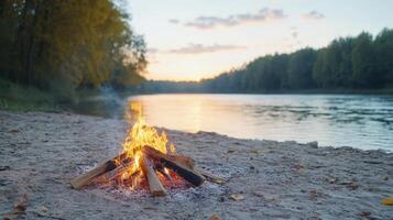 serene campfire burns brightly on sandy riverbank, with beautiful sunset reflecting on water photo
