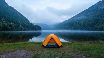 Vibrant orange tent stands beside tranquil lake, surrounded by misty mountains and lush greenery photo