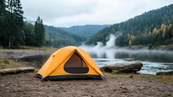Vibrant orange tent is set up beside serene river, surrounded by lush green trees and mist photo