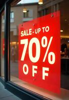 A red sign hanging on a glass door that says SALE - UP TO 70 percent OFF with a blurred background of a store photo
