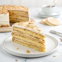 A slice of layered cake with white frosting and nuts on a white plate, a cup of coffee and cookies in the background photo