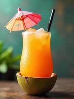 Orange cocktail with ice and a small umbrella on a coconut shell with a straw, green plant in the background photo