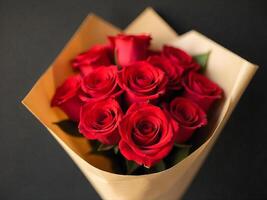 A bouquet of red roses wrapped in brown paper, the roses are in full bloom, with dark green leaves, the background is a dark gray color photo