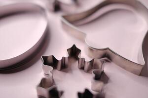 Close-up view of stainless steel cookie cutters in festive shapes including a snowflake, bell, and circle, placed on a soft pink background. photo