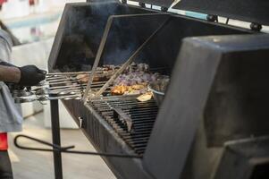 Male chef preparing meat on grill at barbecue party in summer garden, chef's hands in gloves. Concept of food, people and family time. Pork meat on skewers is roasted on coals in large metal grill device photo