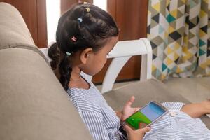 A little Asian girl with ponytail hair sitting on sofa while using smartphone to watch photo