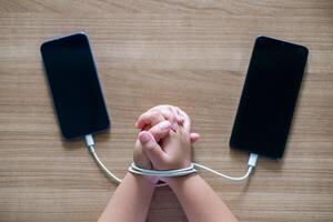 Child's Hands tied with charging cables between two smartphones on wooden table, symbolizing smartphone addiction and dependency in modern digital life. Conceptual image for technology overuse photo