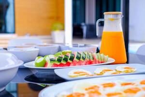 Colorful breakfast setup with sliced watermelon, guava, and cucumber served on a white plate, alongside a jug of fresh orange juice and sunny side up eggs photo
