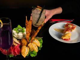 Close-up of a table with two cocktails, a glass of purple drink and a clear mixed drink held by a hand, and plates of various appetizers including skewers and mozzarella. photo