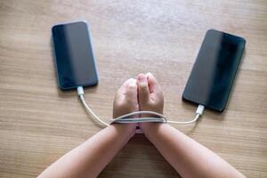 Child's Hands tied with charging cables between two smartphones on wooden table, symbolizing smartphone addiction and dependency in modern digital life. Conceptual image for technology photo