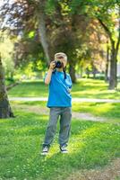 View of cute boy in blue t-shirt taking pictures with camera in park photo