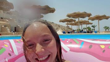alegre Adolescência menina flutuando em Rosa inflável rosquinha anel, espirrando e fazer brincalhão expressões enquanto desfrutando ensolarado recorrer natação piscina durante verão período de férias video