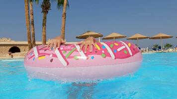 Smiling young girl enjoying summer vacation at resort pool, playing with pink inflatable donut while adjusting swimming goggles and floating carefree video