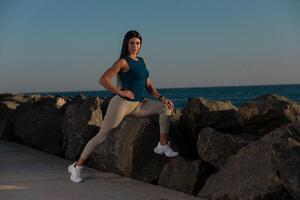 woman performs stretching exercise balancing on rocky shoreline as sun sets in background. ocean waves create serene atmosphere while she focuses on her workout photo
