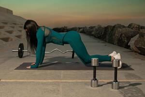 fitness enthusiast dressed in teal activewear performs push up exercises on mat positioned on rocky shoreline. Dumbbells are placed nearby as sun sets creating serene backdrop photo