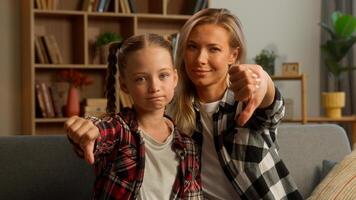 Caucasian family child schoolgirl looking at camera demonstrating thumb down upset little kid mother discontent sad mom woman female showing negative response parent daughter denial no gesture at home photo