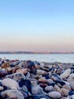 Close-up photo of colorful smooth pebbles on a beach with a blurred background of calm sea and pastel sky. Natural texture and soft focus foreground.