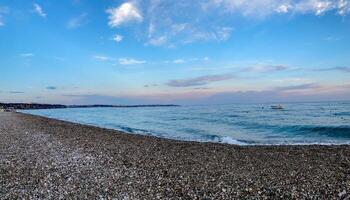Panoramic view of calm sea and pebble beach under a blue sky with pastel sunset clouds and a small boat on the horizon. Peaceful coastal landscape with natural colors. photo