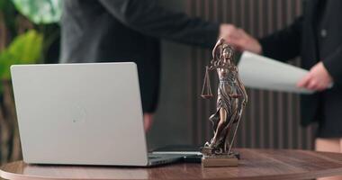 A laptop and a statue of justice against a background of business people shaking hands while discussing a course of action. The concept of legal support and justice video