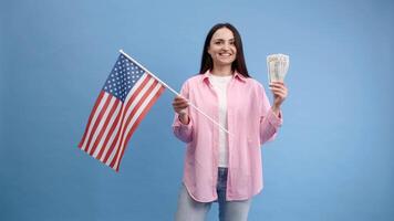 Young brunette woman in pink shirt poses on blue background in studio with american flag and hundred dollar cash with passport. Concept of success, travel, knowledge of foreign language video