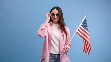 Young smiling woman wears casual clothes holding american flag and looking to the camera isolated on blue color background video