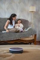 Robot vacuum cleaning the floor while a mother and daughter relax together on the couch in the background. Concept of smart home technology freeing time for family connection. High quality photo