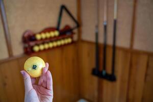 Hand holding billiard ball number 12 in front of a home billiard setup with cues and rack. Concept of indoor recreation and personal hobby space. High quality photo